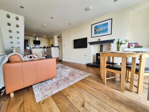 A living room with a sofa, a dining table with chairs, a fireplace, and a kitchen in the background. - 2 Bed in Aberdyfi (oc-dy037) (Aberdovey)