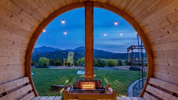 Un sauna en bois, doté de bancs et d’un système de chauffage central, est entouré d’une structure semi-circulaire en bois offrant une vue sur un paysage montagneux.