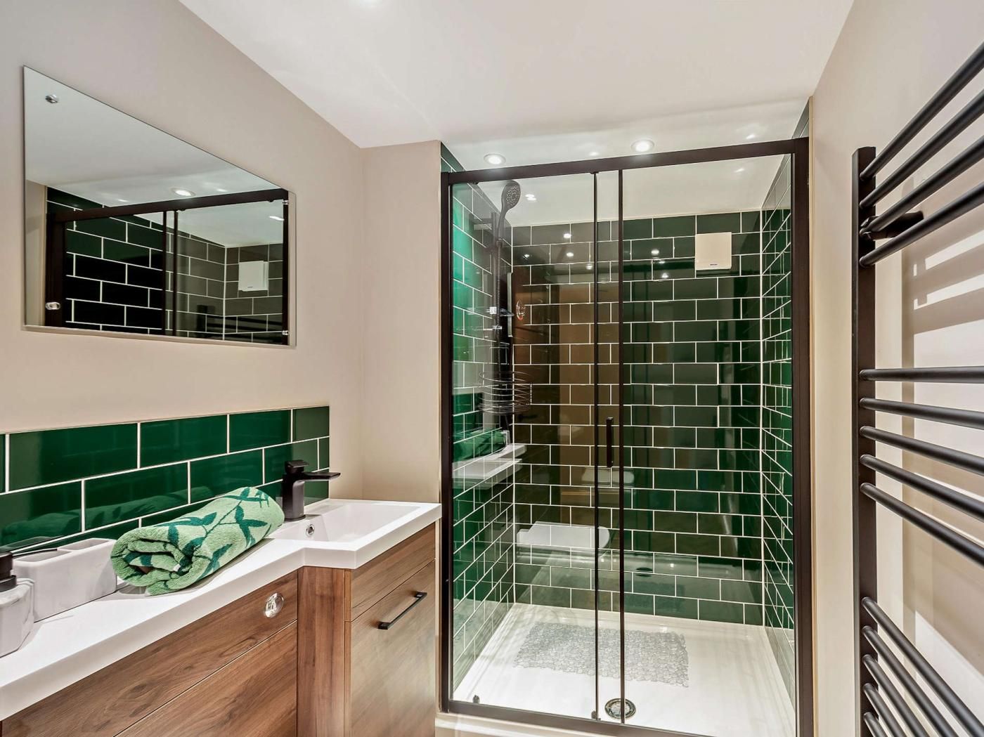 A modern bathroom with a glass shower enclosure, a wooden vanity, and a green-tiled backsplash.