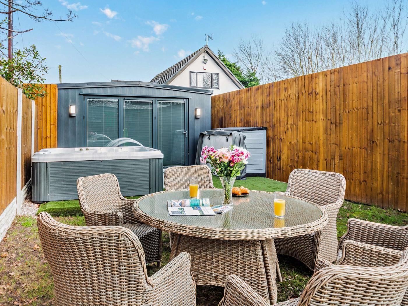 A backyard with a hot tub, wicker chairs, a table with a vase of flowers, and a wooden fence.