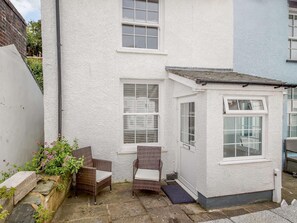 A small, white-walled building with two windows and a door, a small porch area with wicker chairs, and a stone pathway. - 1 Bed in Aberdovey (oc-87910) (Aberdovey)