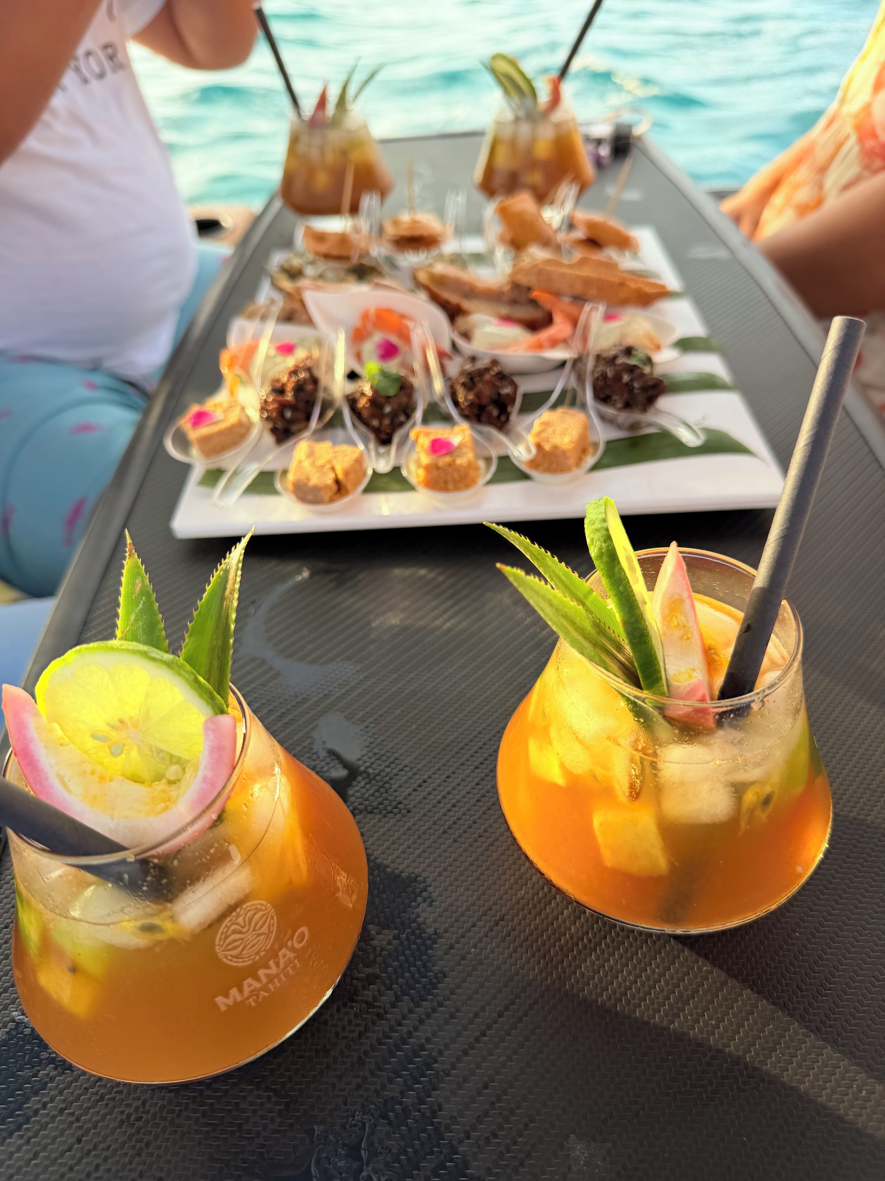 A table with drinks garnished with lime and aloe vera, and a variety of desserts on a white plate.