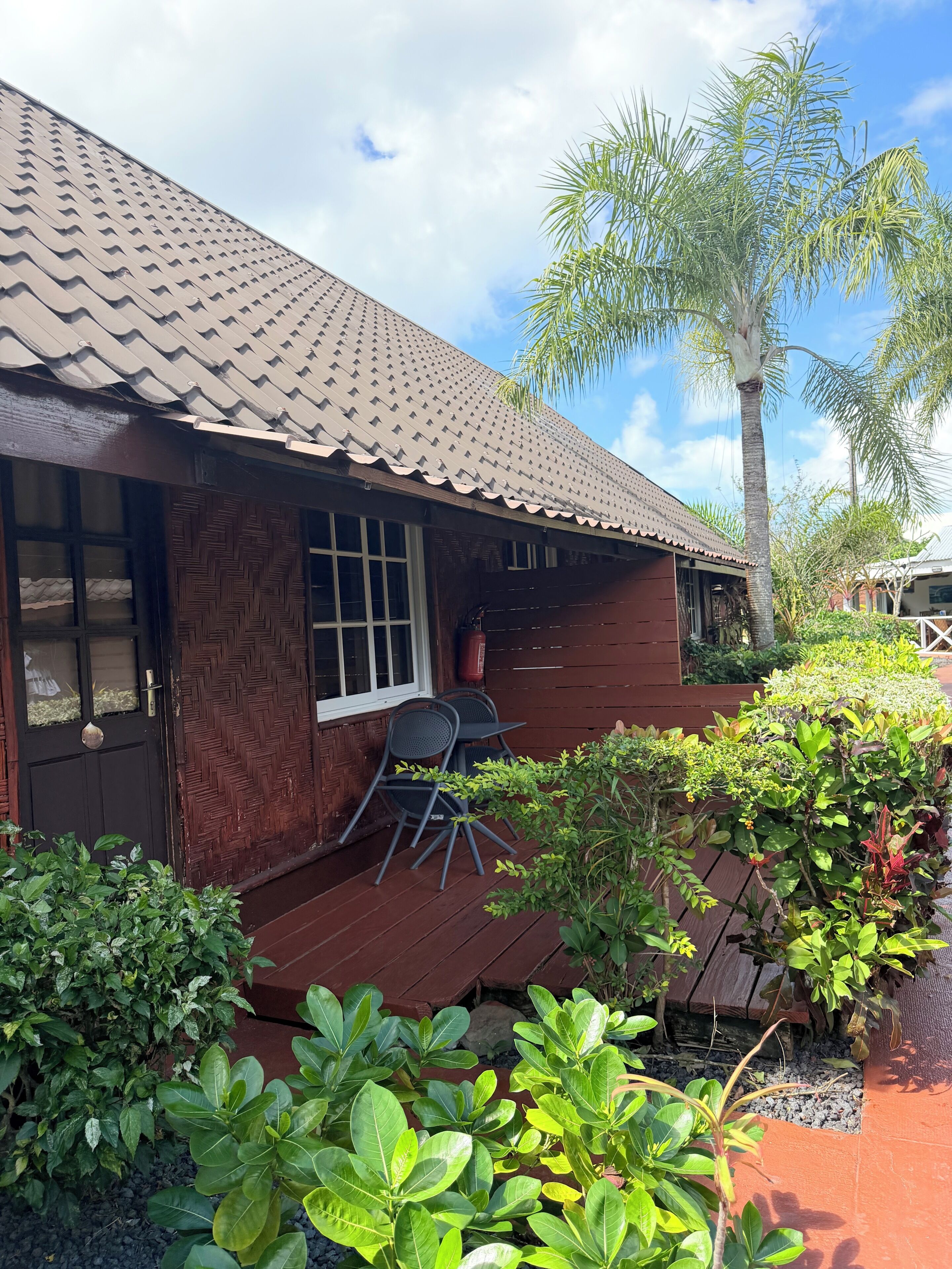 A wooden house with a tiled roof, a porch with chairs, and lush greenery.