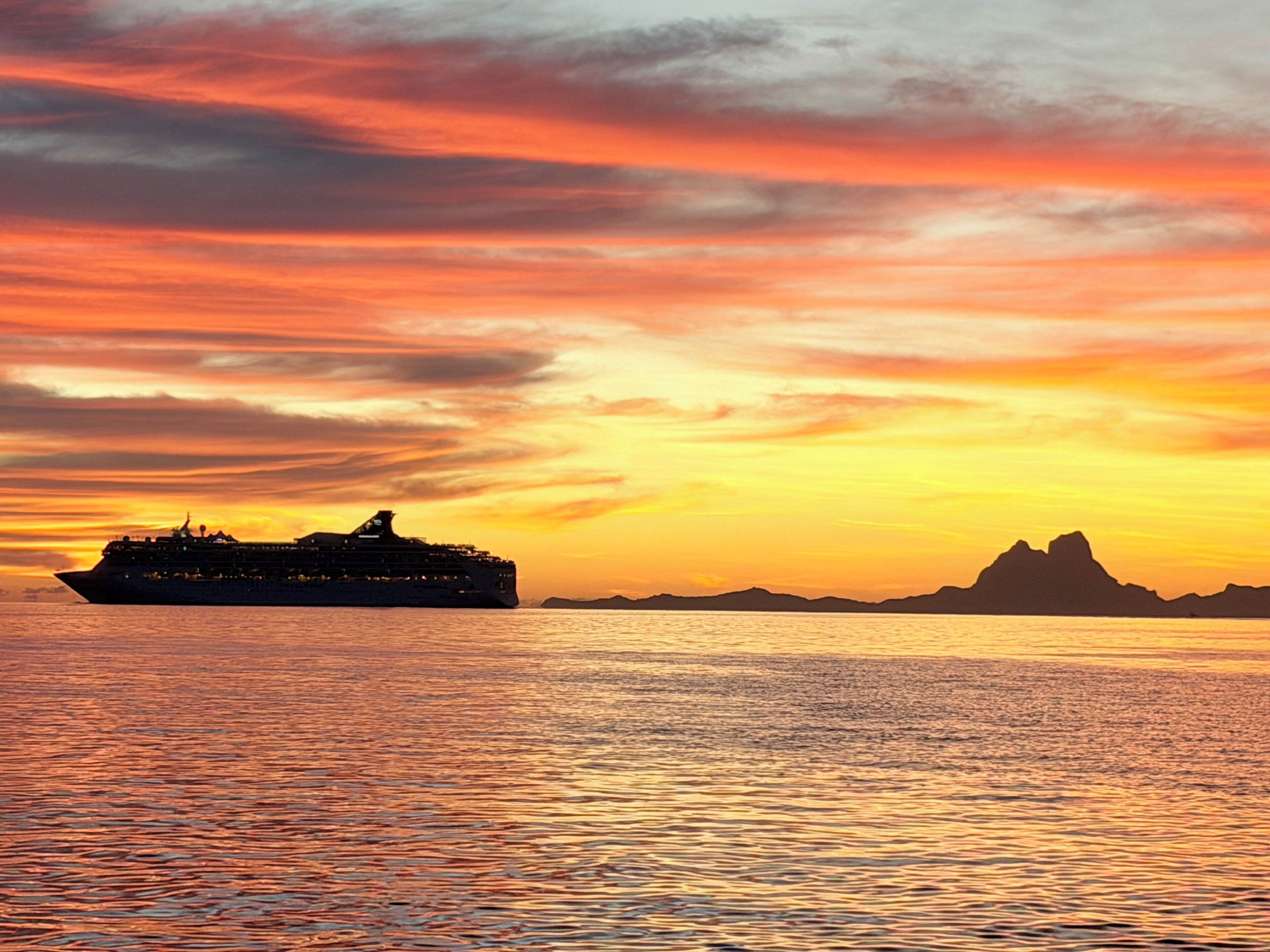 A cruise ship at sunset with a mountainous coastline in the background.