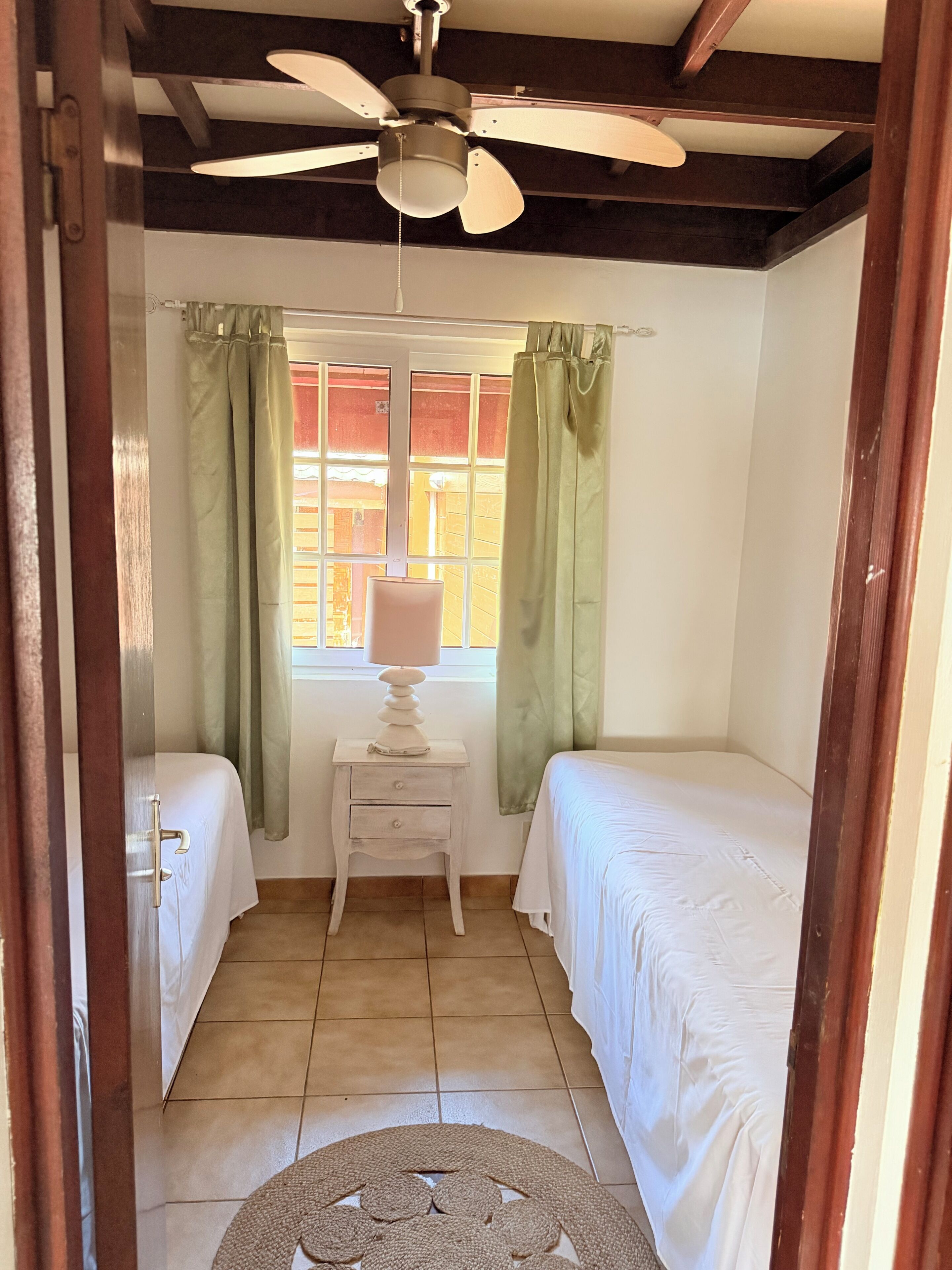 A room with two beds, a ceiling fan, a window with curtains, and a bedside table with a lamp.