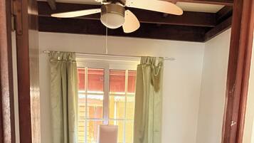 A room with two beds, a ceiling fan, a window with curtains, and a bedside table with a lamp.