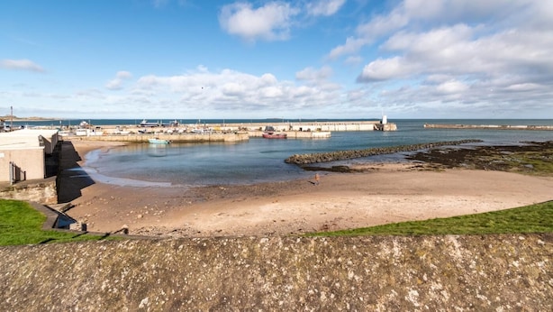 Eine Küstenlandschaft mit einem Sandstrand, einem Steg mit Booten und einem Leuchtturm in der Ferne.