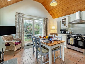 A dining area with a table set for two, a wicker chair, a television, and a kitchen with a stove and oven. - 1 Bed in Keswick (oc-94245) (Keswick)