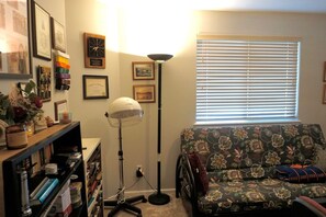 Ein Zimmer mit einer Couch, einer Stehlampe, einem Bücherregal und einem Fenster mit Jalousien.