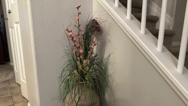 Un angolo con un vaso decorativo grande, pieno di piante verdi e fiori rosa, una porta bianca e una scala con ringhiere bianche.