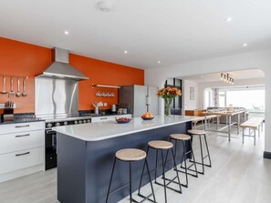 A modern kitchen with a central island, bar stools, and a dining area with a view. - 5 Bed in Penzance (oc-82155) (Gulval)