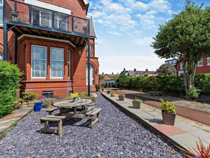 A courtyard with a wooden table and benches, surrounded by potted plants and a gravel path. - 2 Bed in Lytham St. Annes (oc-94212) (Saint Anne's on the Sea)