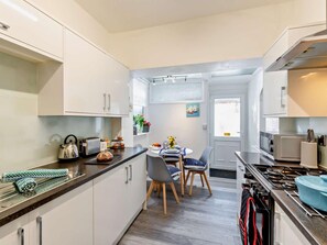 A compact kitchen with white cabinets, a dining area with two chairs, and a view through an open door. - 2 Bed in Lytham St. Annes (oc-94212) (Saint Anne's on the Sea)