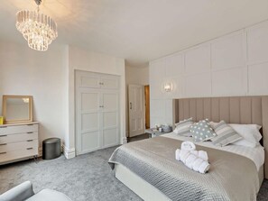 A bedroom with a large bed, a chest of drawers, a mirror, a chandelier, and a door leading to another room. - 2 Bed in Lytham St. Annes (oc-94212) (Saint Anne's on the Sea)