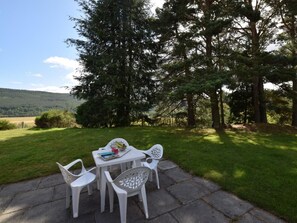 Uma área externa com mesa e cadeiras, rodeada por um gramado e árvores.