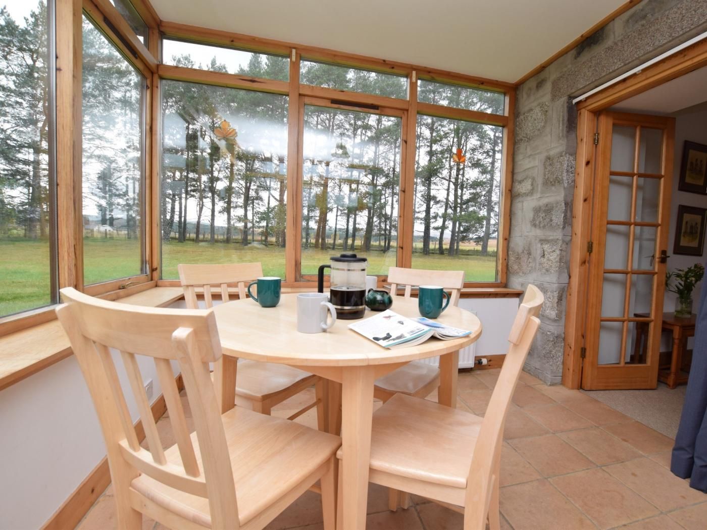 Una mesa de comedor de madera con sillas, una cafetera y un libro en una habitación con ventanas amplias que dan a un bosque.