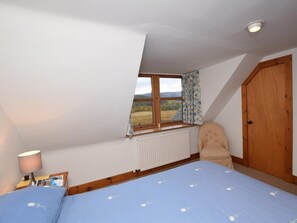 Quarto com teto inclinado, janela com vista para um campo, radiador, cama com colcha florida azul, uma cadeira e uma porta de madeira.
