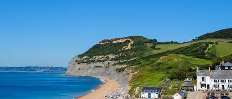 A coastal hotel with a beach, a hill with greenery, and a clear blue sky.
