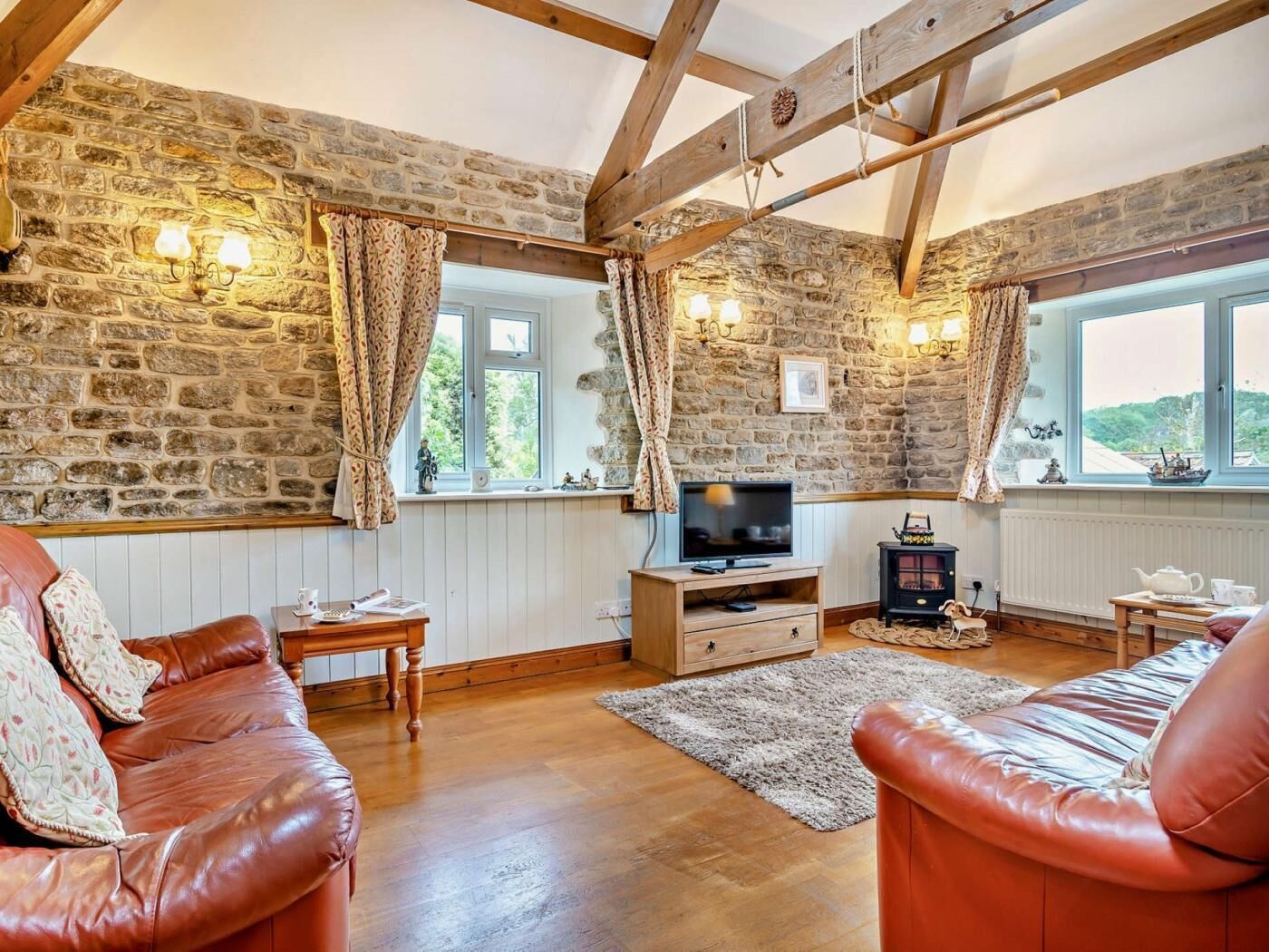A cozy living room with stone walls, wooden beams, a television, a fireplace, and leather sofas.