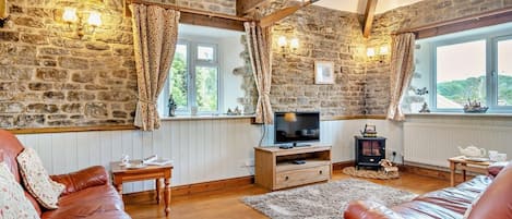 A cozy living room with stone walls, wooden beams, a television, a fireplace, and leather sofas.