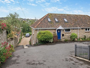 A stone cottage with a blue door, a car parked in the driveway, and a garden with flowering plants. - 2 Bed in Bridport (oc-94222) (Bothenhampton)