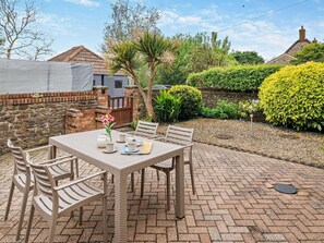 A patio with a table set for four, surrounded by chairs, a brick wall, and a garden with various plants and a hedge. - 2 Bed in Bridport (oc-94222) (Bothenhampton)