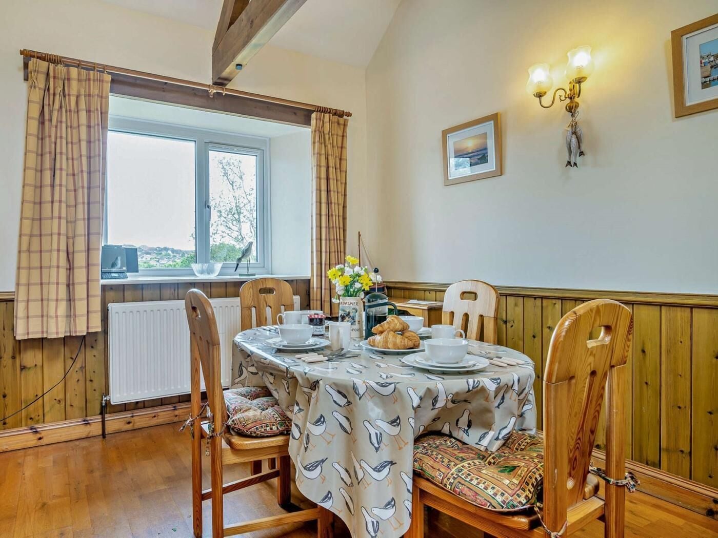 Zona pranzo con tavolo imbandito per quattro persone, sedie in legno, finestra con tende e lampada a parete.