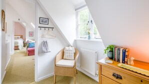Un dormitorio con techo inclinado, una silla de mimbre, un radiador, un aparador con libros y una ventana con vistas.