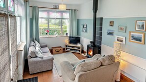 A cozy living room with a fireplace, sofas, a television, and a large window with curtains. - 1 Bed in Redruth (oc-90897) (Redruth)