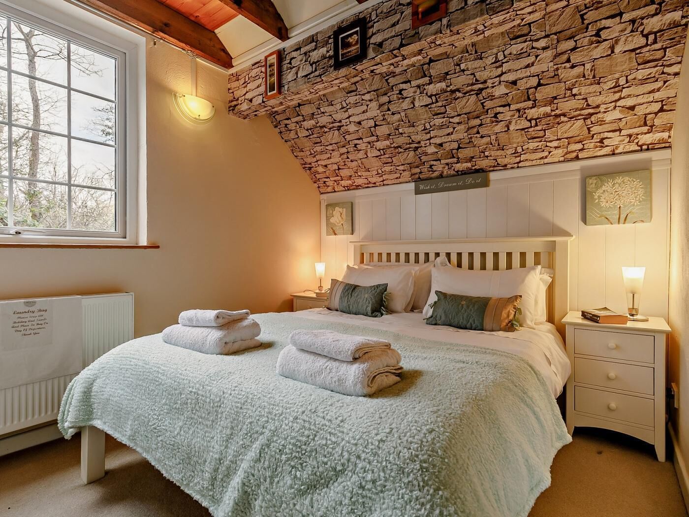 A bedroom with a stone wall, a bed with white bedding, two bedside tables, and a window.