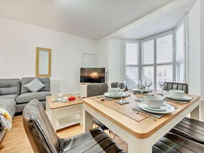 Ein modernes Wohnzimmer mit einem grauen Sofa, einem Holzespeisetisch für vier Personen, einem Fernseher und einem großen Fenster mit Jalousien.