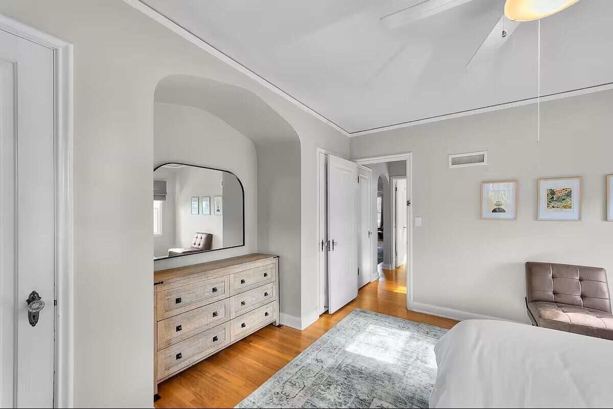 Quarto com cama, cômoda, espelho, uma cadeira e corredor que dá para outro quarto.