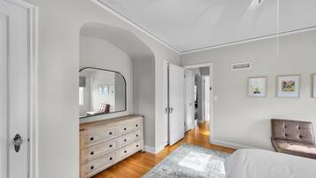 Quarto com cama, cômoda, espelho, uma cadeira e corredor que dá para outro quarto.