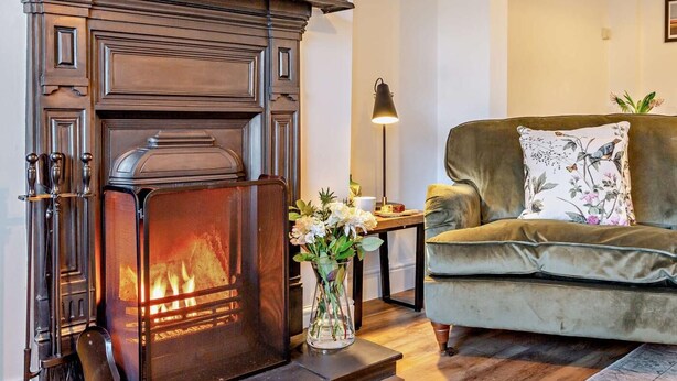 A cozy living room with a wood-burning fireplace, a green sofa, and a vase with flowers. - 2 Bed in Middlesbrough (oc-94220) (Great Broughton)