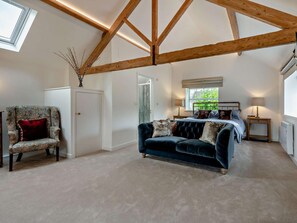 A bedroom with a bed, a dark blue sofa, a floral armchair, and a wooden ceiling structure. - 1 Bed in Blandford Forum (oc-94279) (Okeford Fitzpaine)