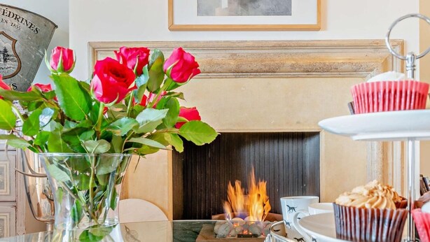 A dining area with a fireplace, a vase of red roses, and a tiered dessert stand. - 4 Bed in Bognor Regis (oc-81446) (Bognor Regis)