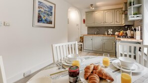 Un comedor con una mesa dispuesta para el desayuno, que incluye croissants, jugo y un frasco de mermelada. Se observa una cocina al fondo, con armarios de colores claros y un salpicadero de azulejos.