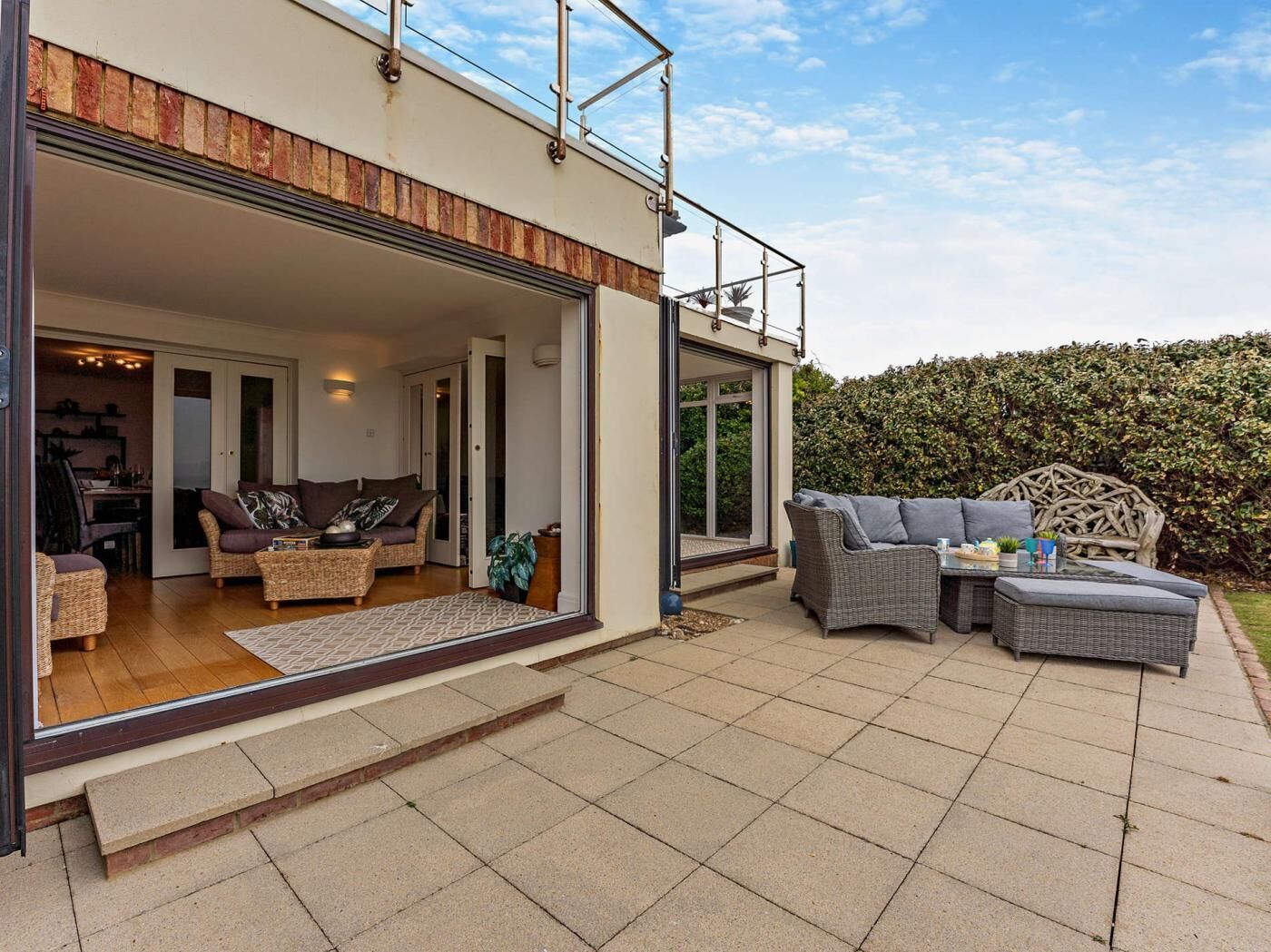 Een modern huis met een terras, rieten meubilair, een eethoek en een grote haag.