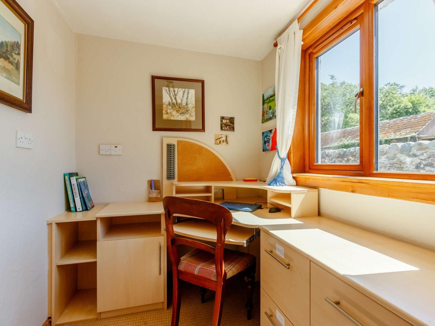 Habitació amb escriptori, cadira, llibres i finestra amb vista a vegetació.
