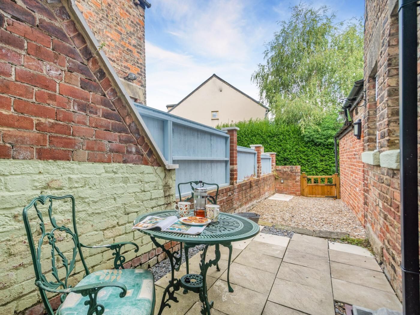 A patio with a table and chairs, a brick wall, a blue fence, and a hedge in the background.