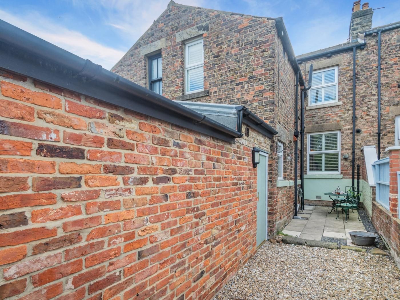 A narrow alleyway with a brick wall on one side and a building with a sloped roof on the other. There is a small outdoor seating area with a table and chairs.