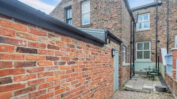 A narrow alleyway with a brick wall on one side and a building with a sloped roof on the other. There is a small outdoor seating area with a table and chairs.