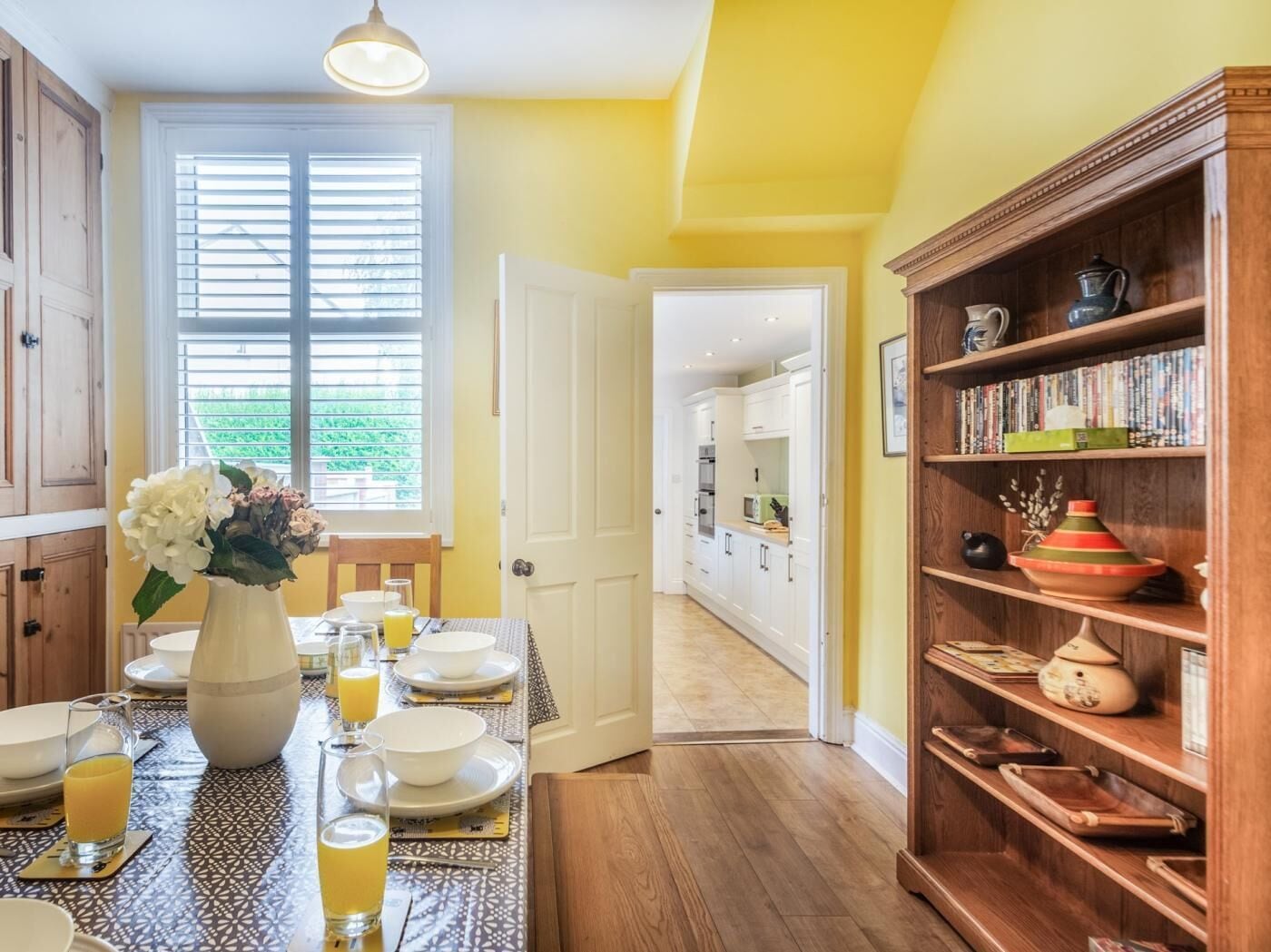 A dining area with a table set for four, a vase of flowers, and a bookshelf with various items.