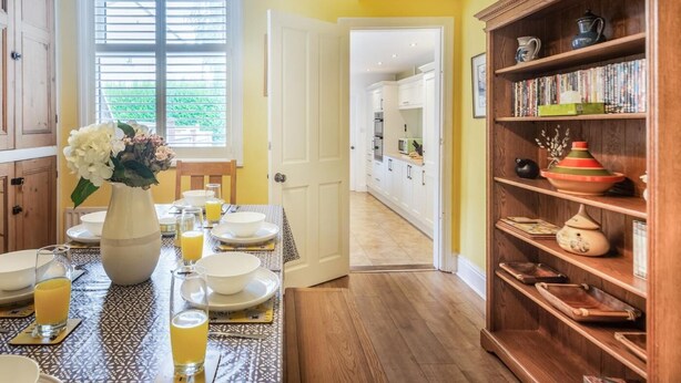 Un comedor con una mesa dispuesta para cuatro, un florero y una estantería con diversos objetos.