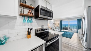 Uma cozinha moderna com micro-ondas, fogão e forno embutidos, uma área de jantar com tapete azul e uma varanda com vista para o oceano.