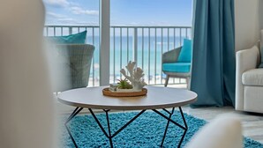 Uma mesa redonda com superfície branca e base trançada, um tapete azul e vista para o oceano através de uma varanda.