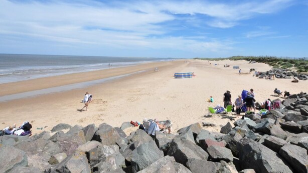 Une plage avec de gros rochers au premier plan, une étendue de sable et un ciel dégagé.