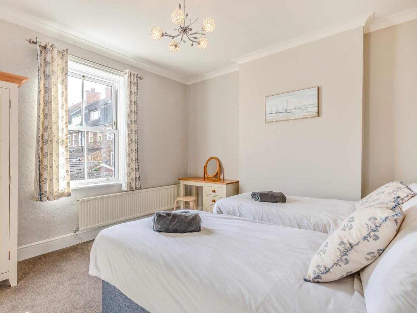 A bedroom with two beds, a dresser, a chair, a window with curtains, a radiator, and a chandelier.