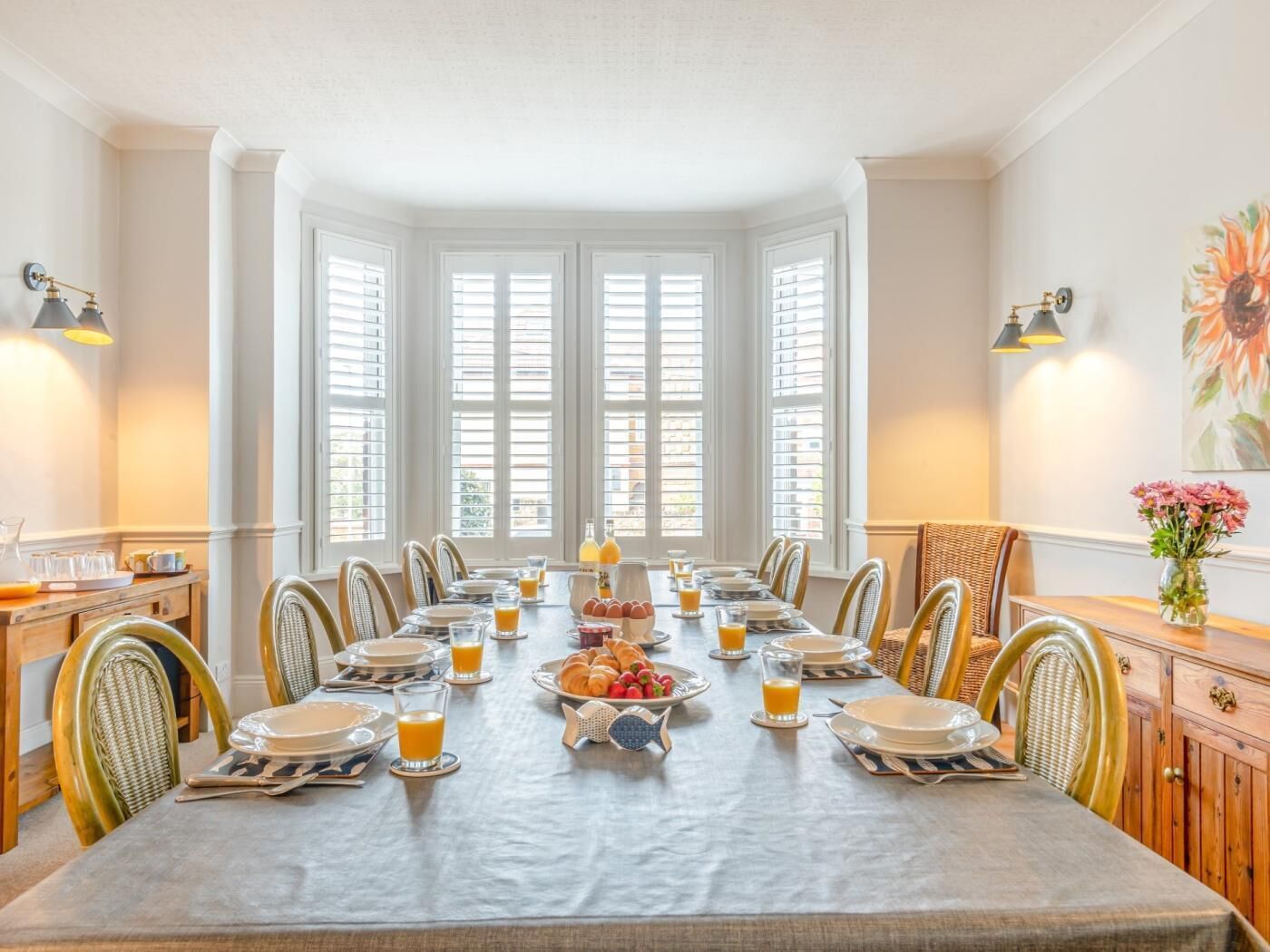 Une salle à manger avec une grande table dressée pour le petit-déjeuner, entourée de chaises en osier, avec une baie vitrée et un tableau floral accroché au mur.