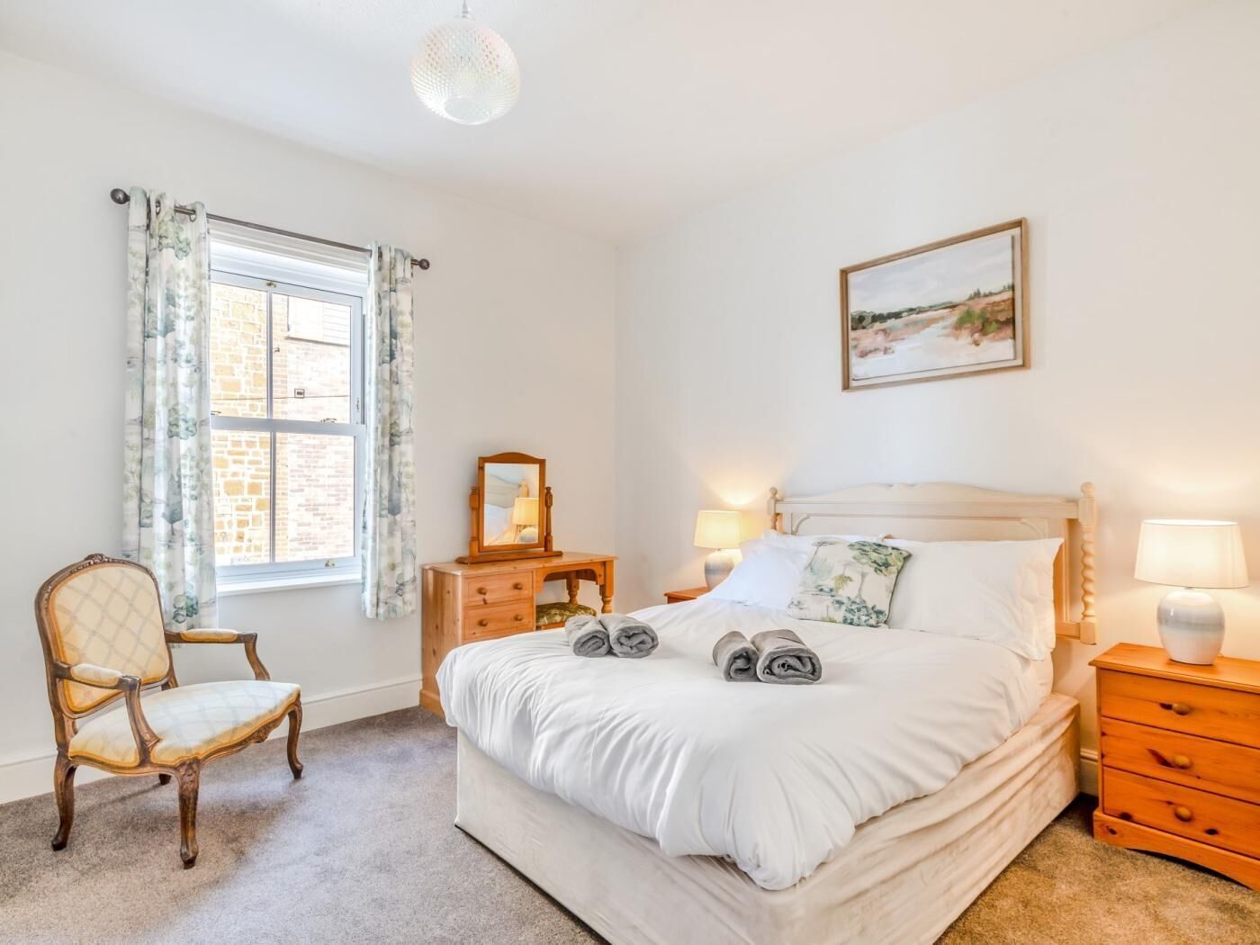 Une chambre à coucher comprenant un lit, une chaise, une commode, une table de chevet, une lampe, un tableau et une fenêtre avec des rideaux.